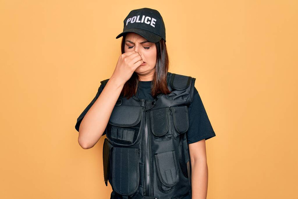 Young beautiful brunette policewoman wearing police uniform bulletproof and cap smelling something stinky and disgusting, intolerable smell, holding breath with fingers on nose. Bad smell - Photo, Image