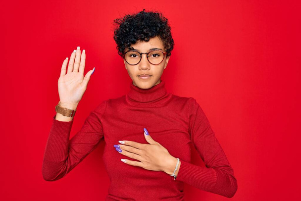 Young beautiful african american afro woman wearing turtleneck sweater and glasses Swearing with hand on chest and open palm, making a loyalty promise oath - Photo, Image