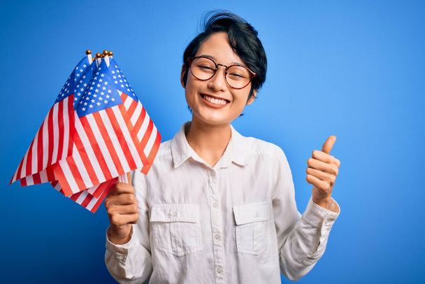 Jeune belle fille patriotique asiatique tenant des drapeaux des États-Unis célébrant le jour de l'indépendance heureux avec un grand sourire faisant signe ok, pouce levé avec les doigts, excellent signe
 - Photo, image