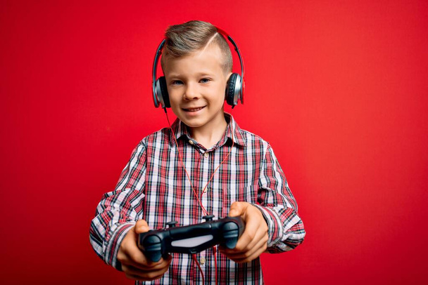 Adorable blond gamer toddler smiling happy and confident. Standing with smile on face playing video game using gamepad and headphones over isolated red background - Foto, Imagem