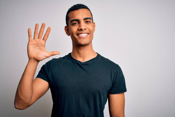 Junger gutaussehender afrikanisch-amerikanischer Mann in lässigem T-Shirt steht vor weißem Hintergrund und zeigt mit Finger Nummer fünf nach oben, während er selbstbewusst und glücklich lächelt. - Foto, Bild