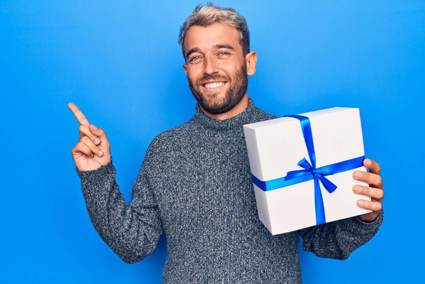Молодой красивый мужчина с бородой, держащий подарок на день рождения на голубом фоне, счастливо улыбается, показывая рукой и пальцем в сторону - Фото, изображение