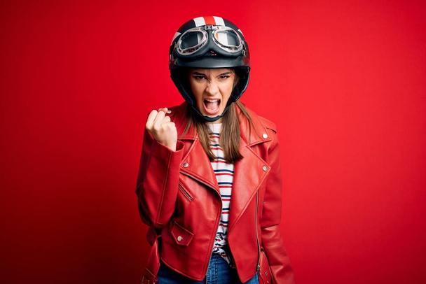 Νεαρή όμορφη μελαχρινή motocyclist γυναίκα φορώντας κράνος μοτοσικλέτας και κόκκινο σακάκι θυμωμένος και τρελός ανύψωση γροθιά απογοητευμένοι και έξαλλος, ενώ φωνάζει με θυμό. Οργή και επιθετική αντίληψη. - Φωτογραφία, εικόνα
