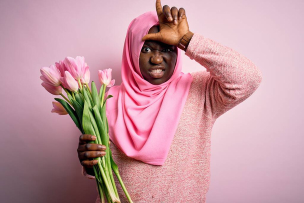 Mladý africký Američan plus velikost žena na sobě muslim hidžáb držení kytice růžové tulipány dělat legraci z lidí s prsty na čele dělá poražený gesto posměch a urážlivé. - Fotografie, Obrázek