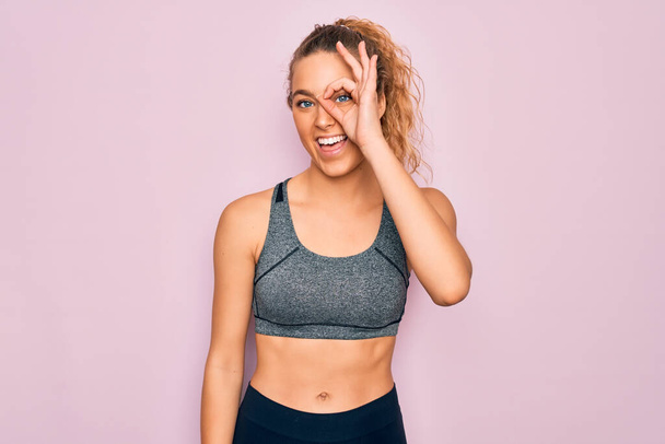Young beautiful blonde sportswoman with blue eyes doing exercise wearing sportswear doing ok gesture with hand smiling, eye looking through fingers with happy face. - Photo, Image