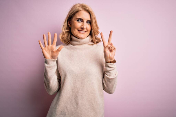 Mujer rubia hermosa de mediana edad con suéter casual cuello alto sobre fondo rosa que muestra y señala con los dedos número siete mientras sonríe confiado y feliz
. - Foto, Imagen