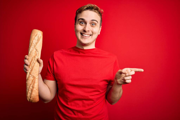 Young handsome redhead man holding fresh homemade bread over isolated red background Smiling happy pointing with hand and finger to the side - Photo, Image
