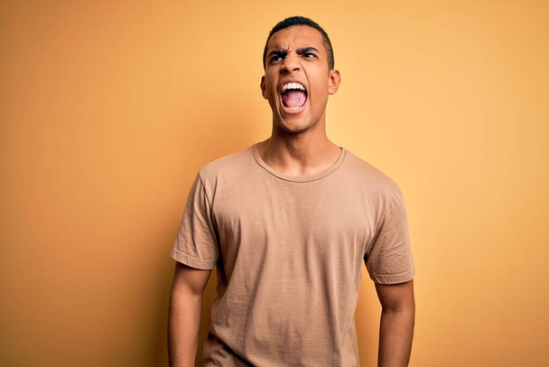 Молодий привабливий афроамериканець у звичайній футболці, що стоїть над жовтим фоном, злий і божевільний, що кричить, розчарований і розлючений, кричить від гніву. Жорстокість і агресивна концепція. - Фото, зображення