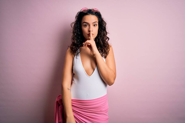 Beautiful woman with curly hair on vacation wearing white swimsuit over pink background asking to be quiet with finger on lips. Silence and secret concept. - Photo, Image
