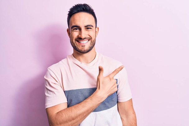 Молодий красивий чоловік з бородою в повсякденній футболці стоїть над рожевим тлом усміхнений веселий вказує рукою і пальцем в сторону
 - Фото, зображення
