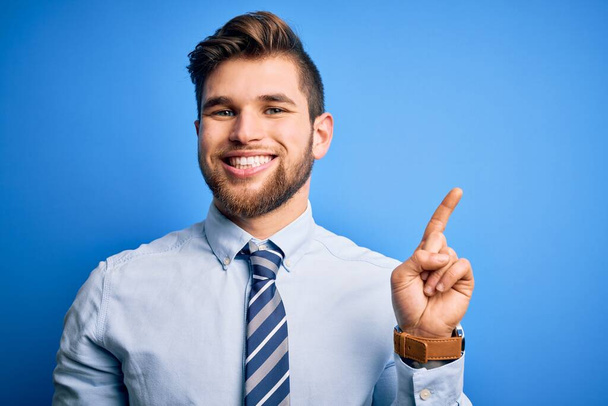 Νεαρός ξανθός επιχειρηματίας με γενειάδα και μπλε μάτια φοράει κομψό πουκάμισο και γραβάτα στέκεται δείχνοντας και δείχνοντας προς τα πάνω με το δάχτυλο νούμερο ένα, ενώ χαμογελά αυτοπεποίθηση και χαρούμενος. - Φωτογραφία, εικόνα