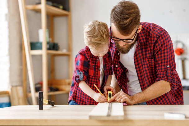 Delighted little boy in casual clothes laughing and helping father to   assembling furniture  and measure the board on table in home workshop togethe - Foto, Imagem