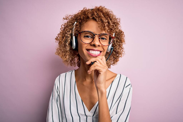 Αφρικανική αμερικανική σγουρή γυναίκα πράκτορας που εργάζεται χρησιμοποιώντας ακουστικά πάνω από ροζ φόντο αναζητούν αυτοπεποίθηση στην κάμερα με χαμόγελο με σταυρωμένα χέρια και το χέρι έθεσε στο πηγούνι. Θετική σκέψη. - Φωτογραφία, εικόνα