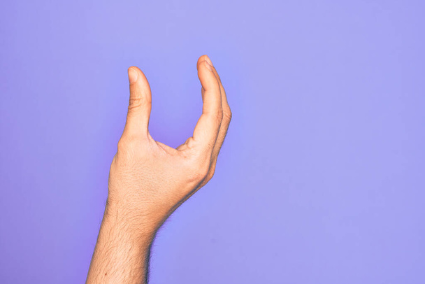 Ręka białego młodzieńca pokazuje palce nad odizolowanym fioletowym podkładem i bierze niewidzialną rzecz, trzymając obiekt z palcami pokazującymi przestrzeń - Zdjęcie, obraz