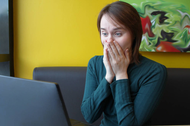 Girl with a perplexed surprised tense expression sits sitting working at a laptop at a table in a cafe on a brown sofa against a yellow wall. She put her hands on face, covered mouth with palms - 写真・画像
