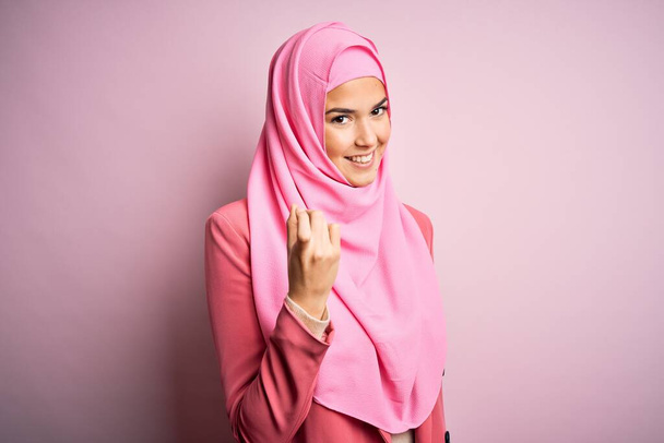 Una guapa jovencita que lleva hijab musulmán de pie sobre fondo rosa aislado Beckoning venir aquí gesto con la mano invitando a la bienvenida feliz y sonriente. - Foto, imagen