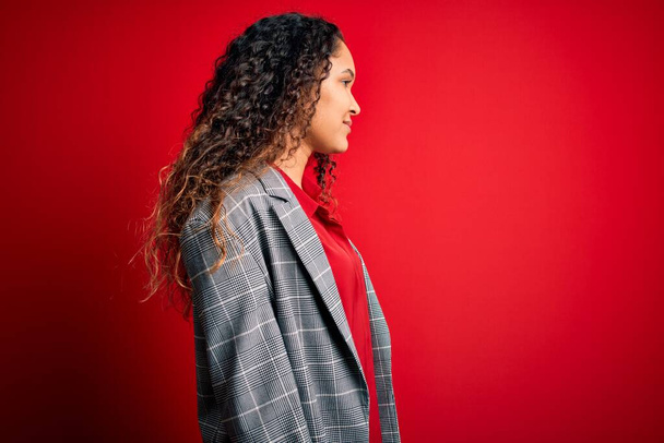 Jovem mulher de negócios bonita com cabelo encaracolado vestindo jaqueta elegante sobre fundo vermelho olhando para o lado, relaxar pose perfil com rosto natural com sorriso confiante
. - Foto, Imagem