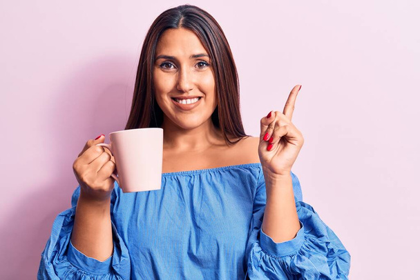Young beautiful brunette woman drinking mug of coffee smiling happy pointing with hand and finger to the side  - Photo, Image