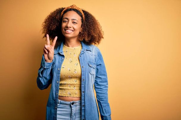 Beautiful african american with afro hair standing over yellow isolated background smiling with happy face winking at the camera doing victory sign. Number two. - Foto, Imagem