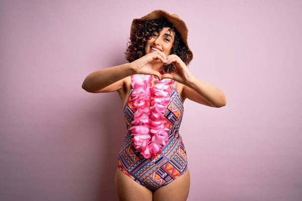 Jonge mooie arabische vrouw op vakantie in badpak en Hawaïaanse lei bloemen glimlachend in liefde doen hart symbool vorm met de handen. Romantisch concept. - Foto, afbeelding