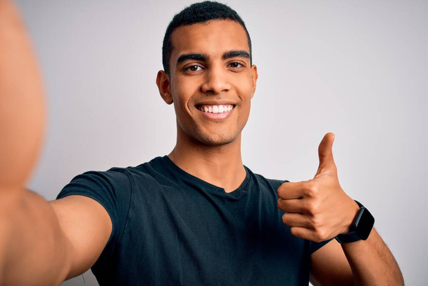 Young handsome african american man wearing casual t-shirt making selfie by camera happy with big smile doing ok sign, thumb up with fingers, excellent sign - Photo, image