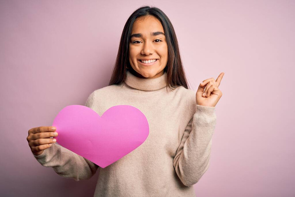 Young beautiful asian woman holding pink heart standing over isolated background very happy pointing with hand and finger to the side - Photo, Image