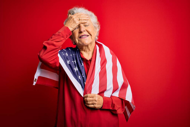 Sênior bela mulher patriótica de cabelos grisalhos vestindo bandeira estados unidos sobre fundo vermelho estressado com mão na cabeça, chocado com vergonha e rosto surpresa, irritado e frustrado. Medo e chateado por engano. - Foto, Imagem