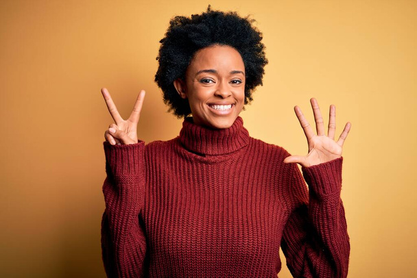 Junge schöne Afro-Amerikanerin mit lockigem Haar trägt lässigen Rollkragenpullover und zeigt mit Finger Nummer sieben nach oben, während sie selbstbewusst und glücklich lächelt. - Foto, Bild