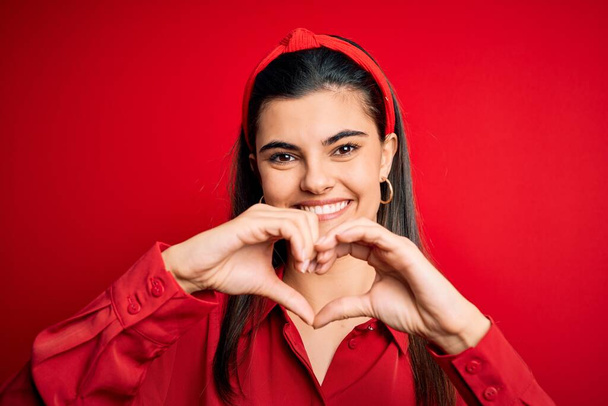 Young beautiful brunette woman wearing casual shirt and diadem over red background smiling in love showing heart symbol and shape with hands. Romantic concept. - Photo, Image