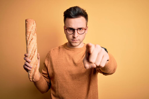 Giovane bell'uomo caucasico con gli occhiali che tiene una fresca baguette di pane puntato con il dito verso la macchina fotografica e verso di voi, segno della mano, gesto positivo e fiducioso dal davanti - Foto, immagini