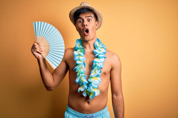 Young tourist man on vacation wearing swimwear and hawaiian lei flowers holding hand fan scared in shock with a surprise face, afraid and excited with fear expression - Foto, imagen