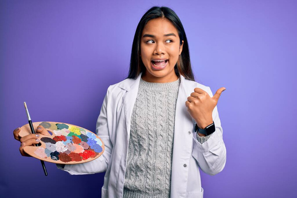 Young asian artist girl painting using paintbrush and palette with colors over purple background pointing and showing with thumb up to the side with happy face smiling - Foto, Imagem
