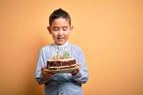Adorable toddler wearing holding birthday cake with candles standing over isolated yellow background - Фото, изображение
