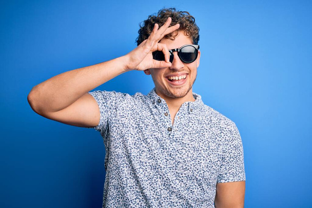 Young blond handsome man with curly hair on vacation wearing striped polo and sunglasses doing ok gesture with hand smiling, eye looking through fingers with happy face. - Photo, Image