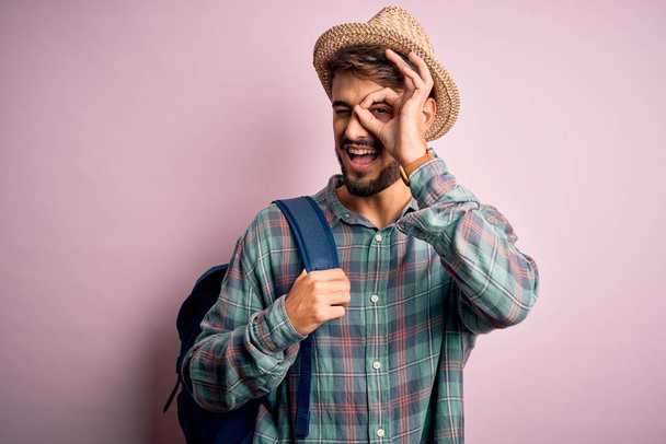 Mladý turista na dovolené na sobě batoh a letní klobouk přes růžové pozadí s veselou tvář s úsměvem dělá ok znamení s rukou na oku dívá skrz prsty - Fotografie, Obrázek