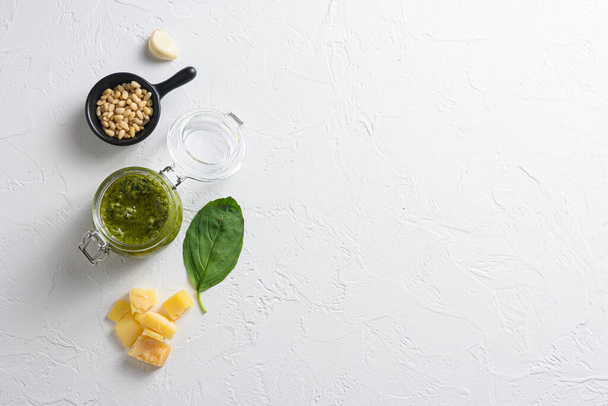 Green Pesto Basil Sauce in glass jar . with ingredients parmesan basil pine nuts on white background top view lay flat space for text. - Photo, Image