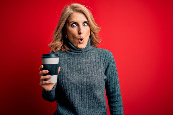 Schöne blonde Frau mittleren Alters trinkt eine Tasse Kaffee zum Mitnehmen auf rotem Hintergrund, erschrocken mit einem überraschenden Gesicht, ängstlich und aufgeregt mit angstvollem Ausdruck - Foto, Bild