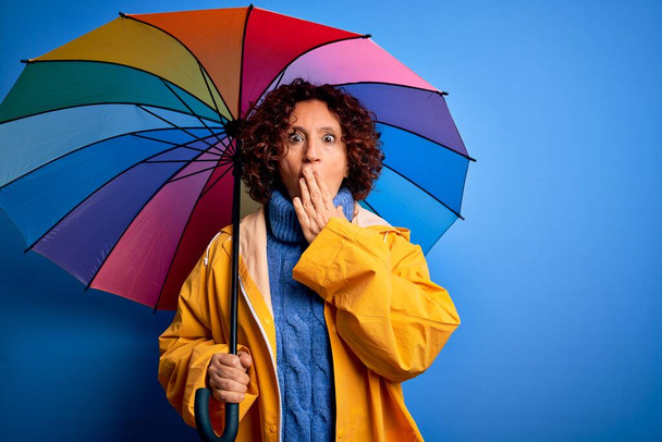 Middle age curly hair woman wearing rain coat holding colorful umbrella over blue background cover mouth with hand shocked with shame for mistake, expression of fear, scared in silence, secret concept - Fotografie, Obrázek