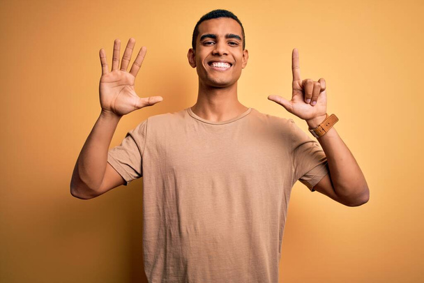 Молодой красивый африканский мужчина в случайной футболке стоит на желтом фоне, показывая и показывая вверх пальцами цифру семь и улыбаясь уверенно и счастливо. - Фото, изображение