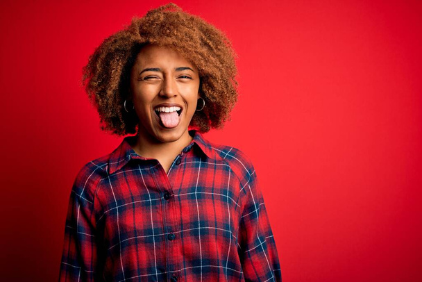 Junge schöne afroamerikanische Afro-Frau mit lockigem Haar in lässigem Hemd, das die Zunge herausstreckt, glücklich mit witzigem Ausdruck. Emotionales Konzept. - Foto, Bild
