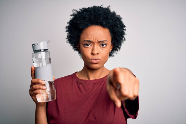 Junge Afro-Amerikanerin mit lockigem Haar, die zur Erfrischung eine Flasche Wasser trinkt und mit dem Finger auf die Kamera und Sie zeigt, Handzeichen, positive und selbstbewusste Geste von vorne - Foto, Bild