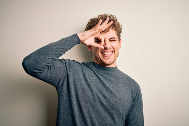 Young blond handsome man with curly hair wearing casual sweater over white background doing ok gesture with hand smiling, eye looking through fingers with happy face. - Photo, Image
