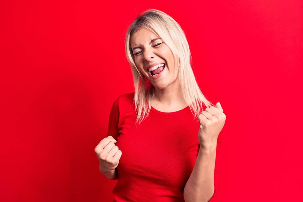 Junge schöne blonde Frau in lässigem rotem T-Shirt steht vor isoliertem Hintergrund und feiert überrascht und erstaunt über den Erfolg mit erhobenen Armen und geschlossenen Augen - Foto, Bild