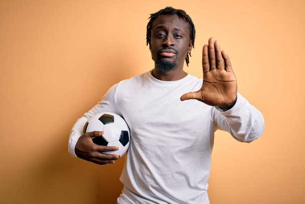 Giovane giocatore africano americano che gioca a calcio tenendo palla da calcio su sfondo giallo con mano aperta facendo stop segno con espressione seria e fiduciosa, gesto di difesa - Foto, immagini
