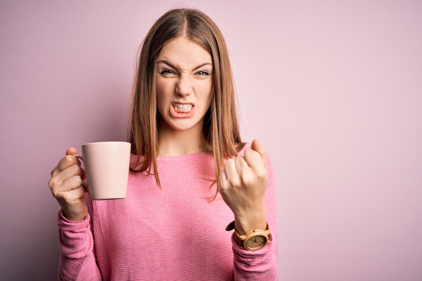 Young beautiful redhead woman drinking mug of coffee over isolated pink background annoyed and frustrated shouting with anger, crazy and yelling with raised hand, anger concept - Zdjęcie, obraz