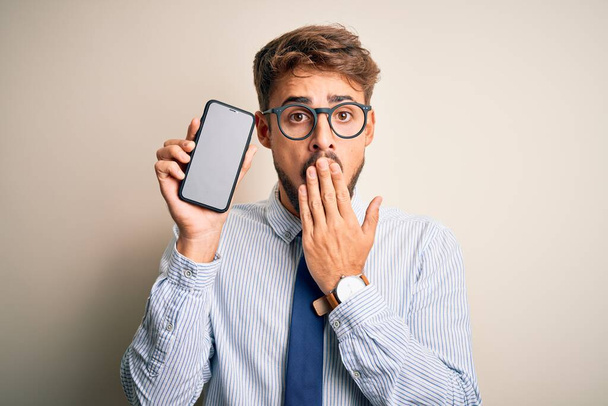 Junger Geschäftsmann mit Smartphone vor isoliertem weißem Hintergrund, der Mund mit der Hand vor Scham über Fehler geschockt, Ausdruck der Angst, Angst in der Stille, Geheimkonzept - Foto, Bild