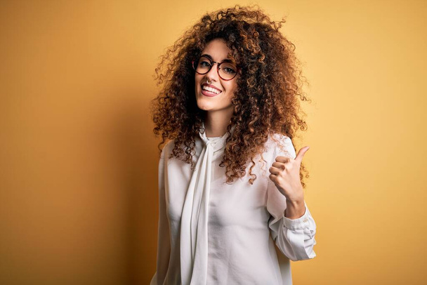 Young beautiful brunette woman with curly hair and piercing wearing shirt and glasses smiling with happy face looking and pointing to the side with thumb up. - Photo, Image