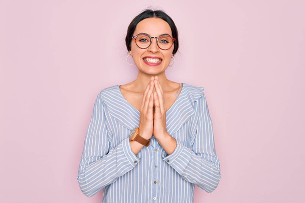 Fiatal gyönyörű nő visel alkalmi csíkos ing és szemüveg felett rózsaszín háttér imádkoznak kéz együtt kér bocsánatot mosolygós magabiztos. - Fotó, kép