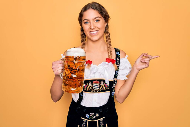 Beautiful blonde german woman with blue eyes wearing octoberfest dress drinking jar of beer very happy pointing with hand and finger to the side - Photo, Image