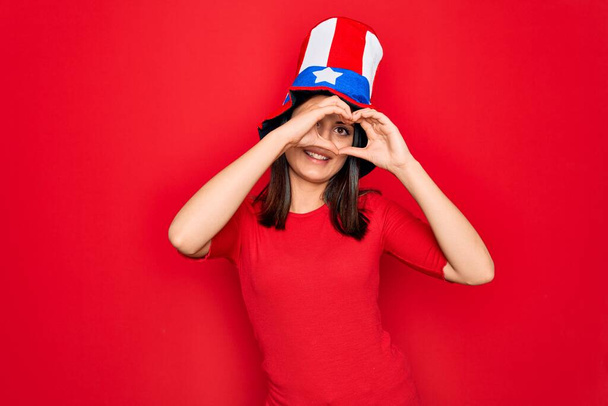 Fiatal gyönyörű barna nő visel egyesült államokbeli kalap ünnepli függetlenség napja csinál szív alakú kéz és ujjak mosolygó keresztül jel - Fotó, kép